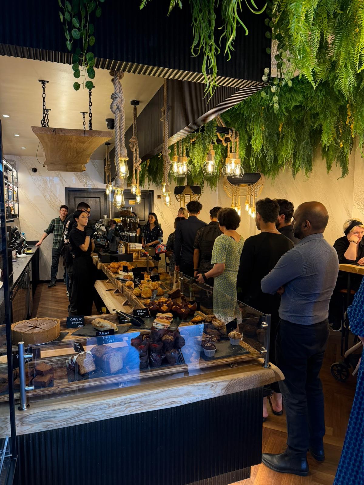 Inside Kappuccino: hanging greenery over a wooden pastry counter with customers queuing
